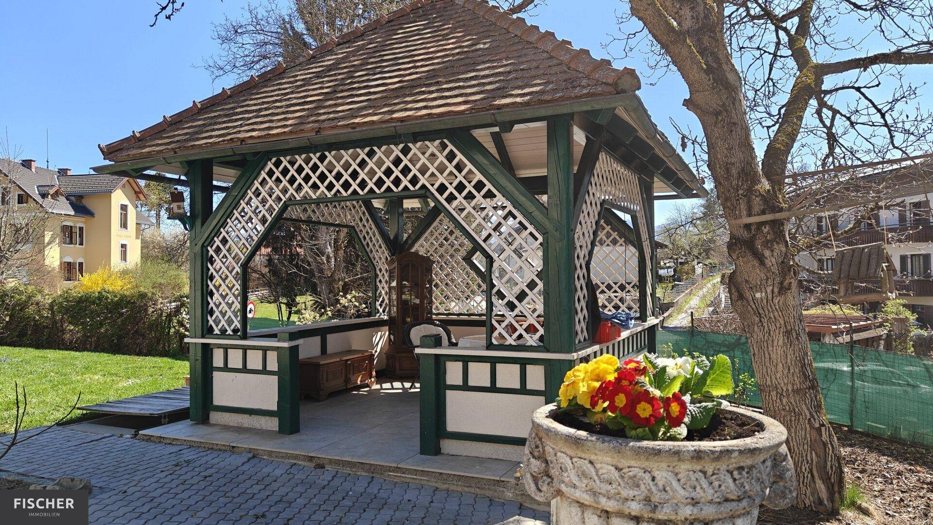 Grüner Garten mit einem dekorativen Holzpavillon und bunten Blumen in einem Steintopf.