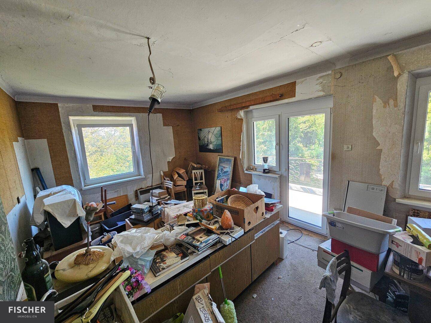 Unaufgeräumter Raum mit mehreren Fenstern und Zugang zu einem Balkon, benötigt Renovierung.