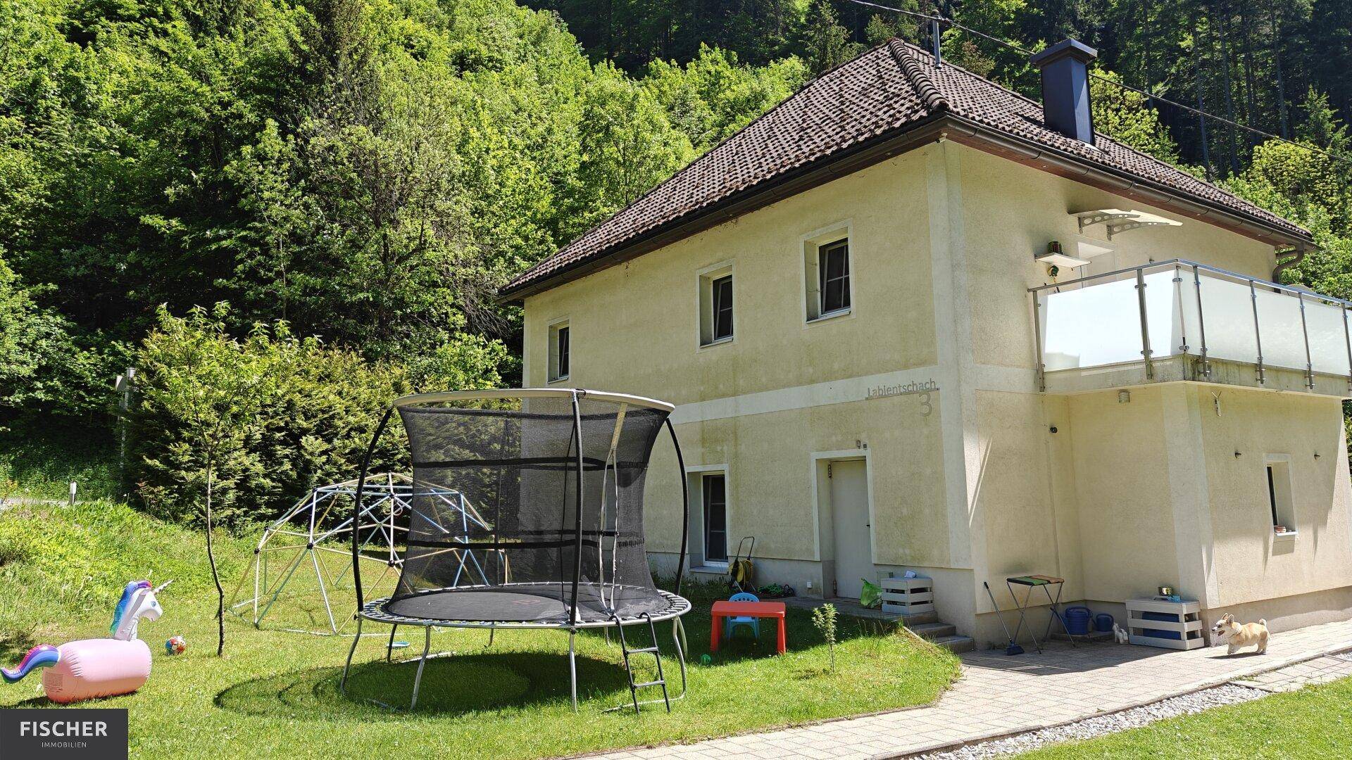 Hausfassade mit großem Garten, Trampolin und Spielgeräten umgeben von viel Grün.