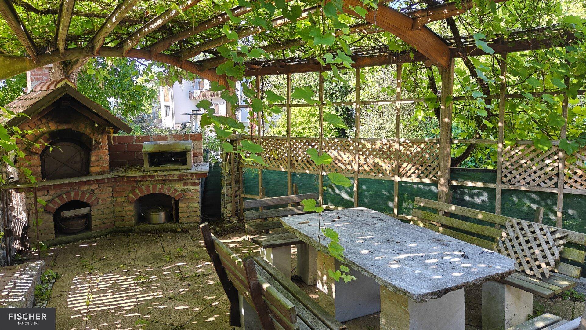 Rustikaler Außenbereich mit gemauertem Pizzaofen, Steintisch und Holzbänken unter einer Pergola.