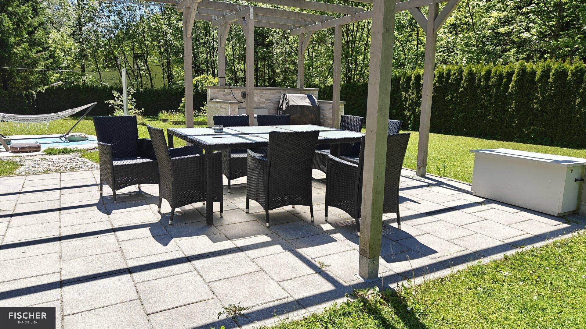 Gepflasterte Terrasse mit großem Esstisch und Stühlen unter einer Holzpergola.
