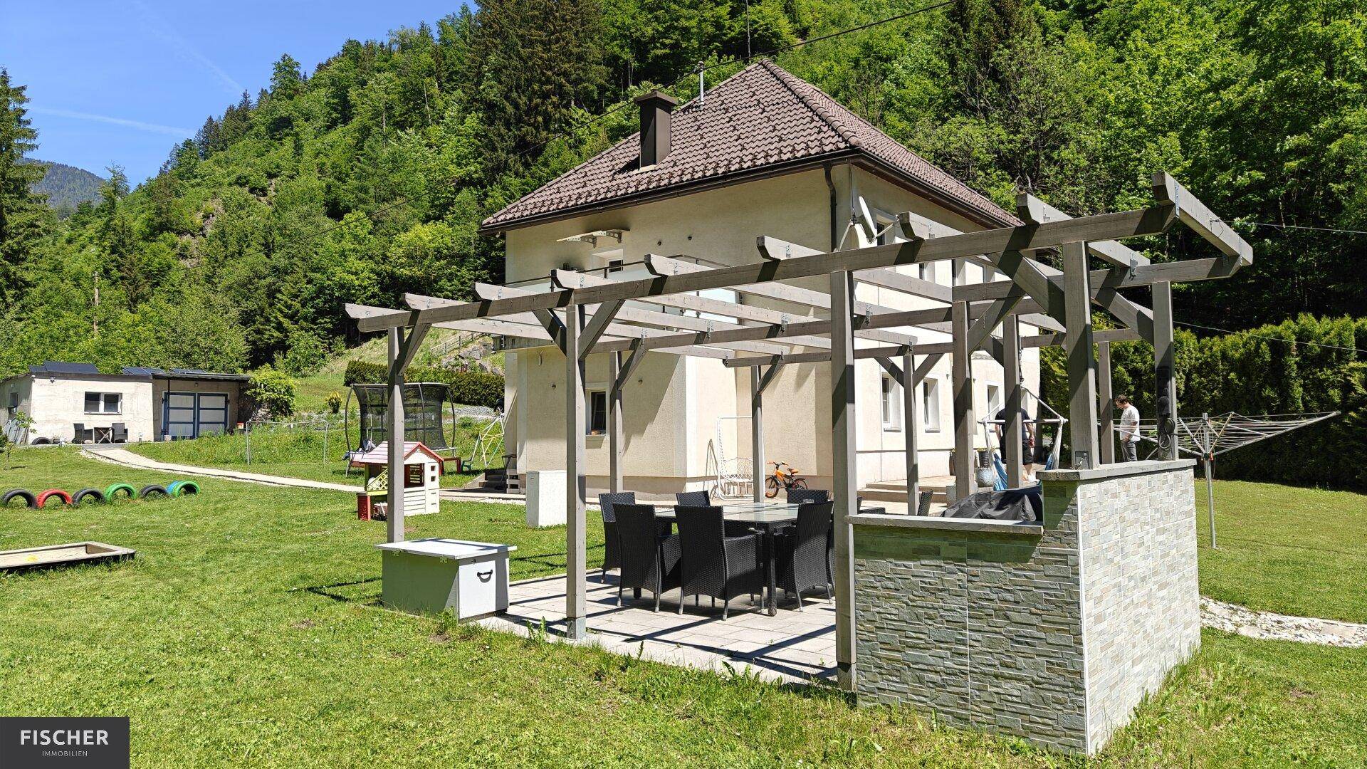 Gartenbereich mit überdachtem Essplatz unter einer Pergola und Blick auf die Natur.