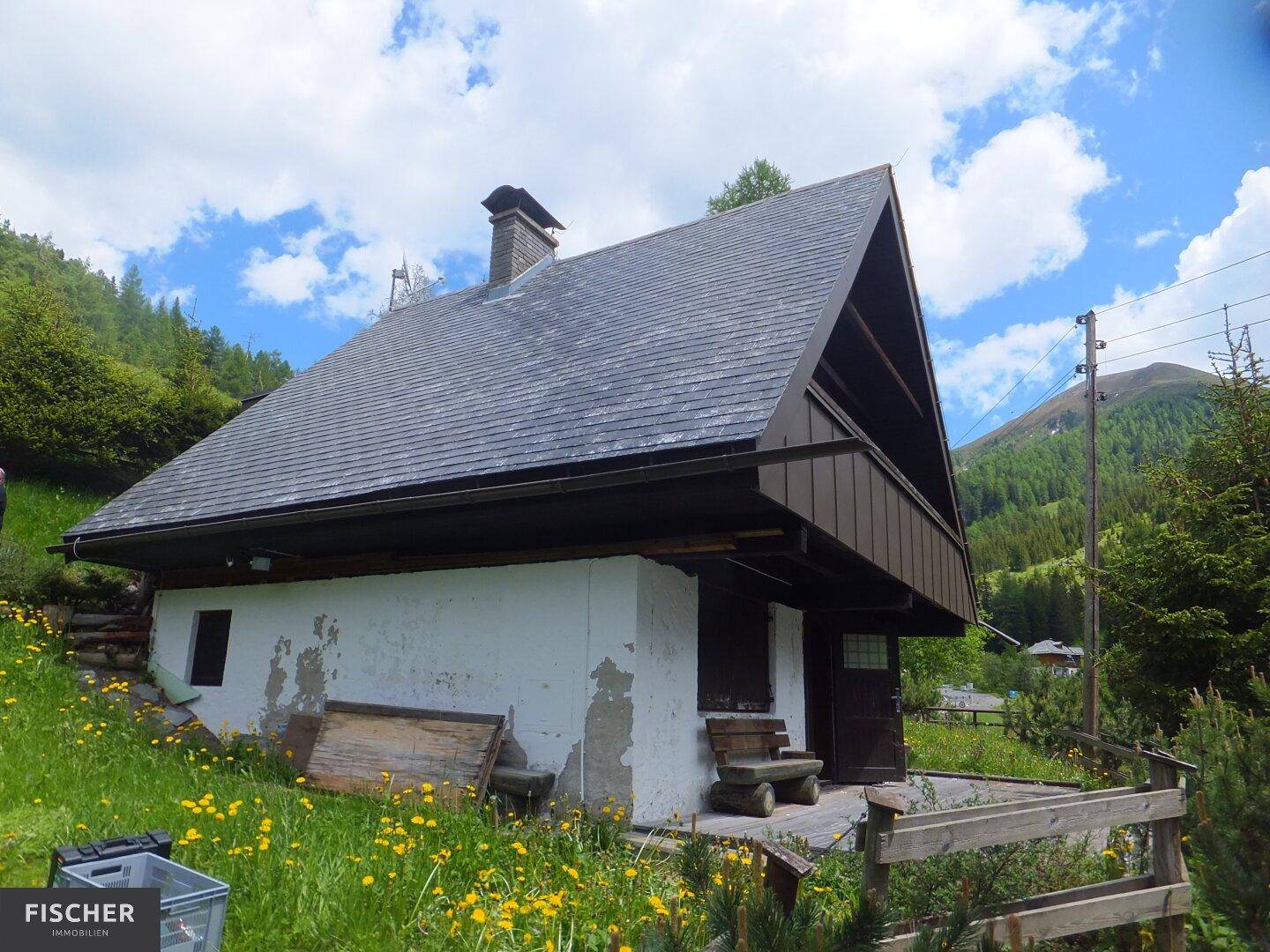 Rustikales Chalet mit Holzdach und weiß getünchter Fassade, umgeben von einer blühenden Wiese in den Bergen.