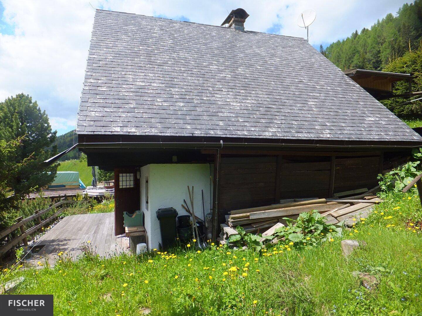 Seitenansicht eines einfachen Holzhauses mit Schindeldach, umgeben von einer grünen Wiese und Bäumen.