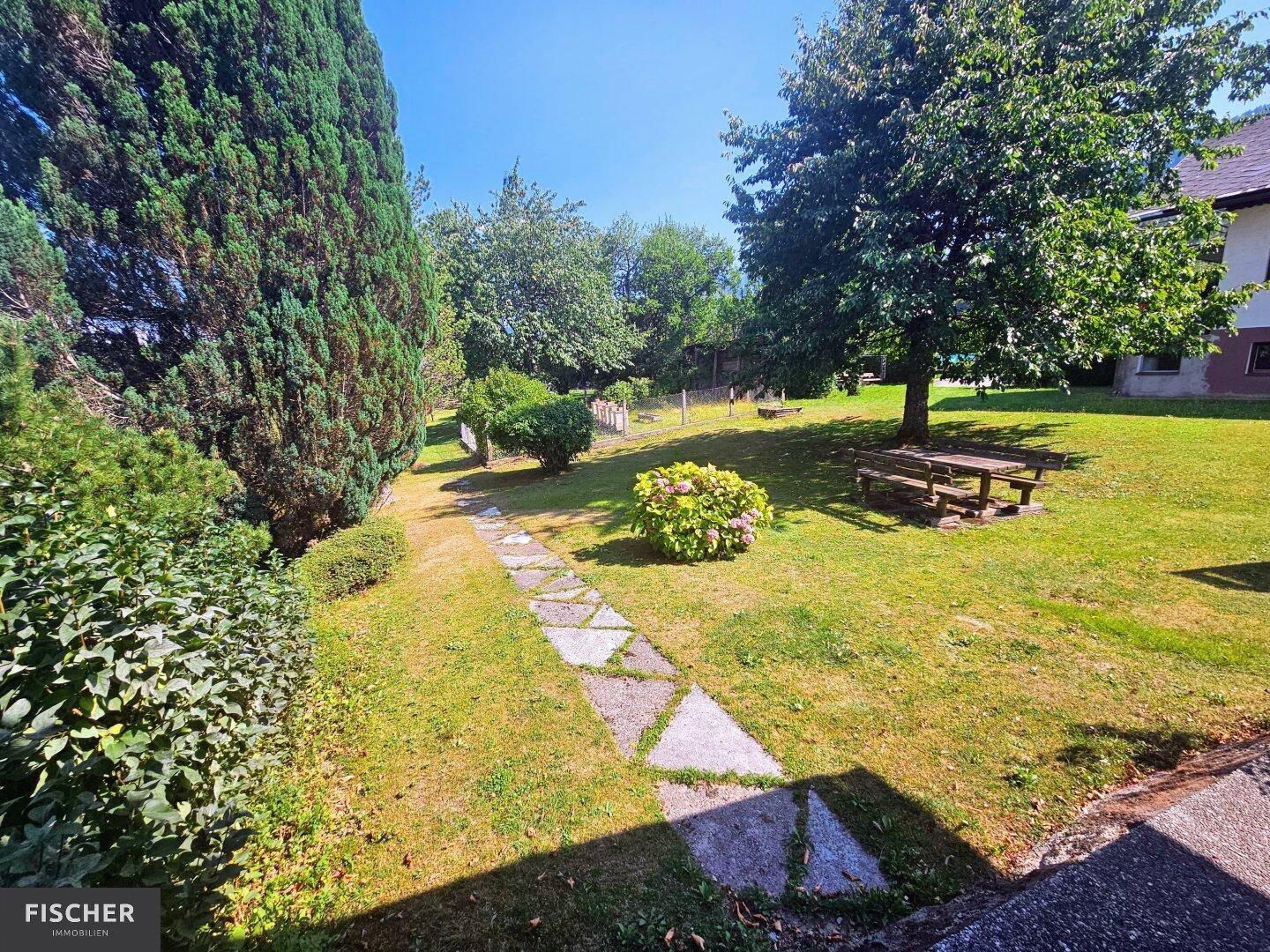 Gepflegter Garten mit Steinweg, üppigem Grün und Sitzbank unter schattenspendenden Bäumen.
