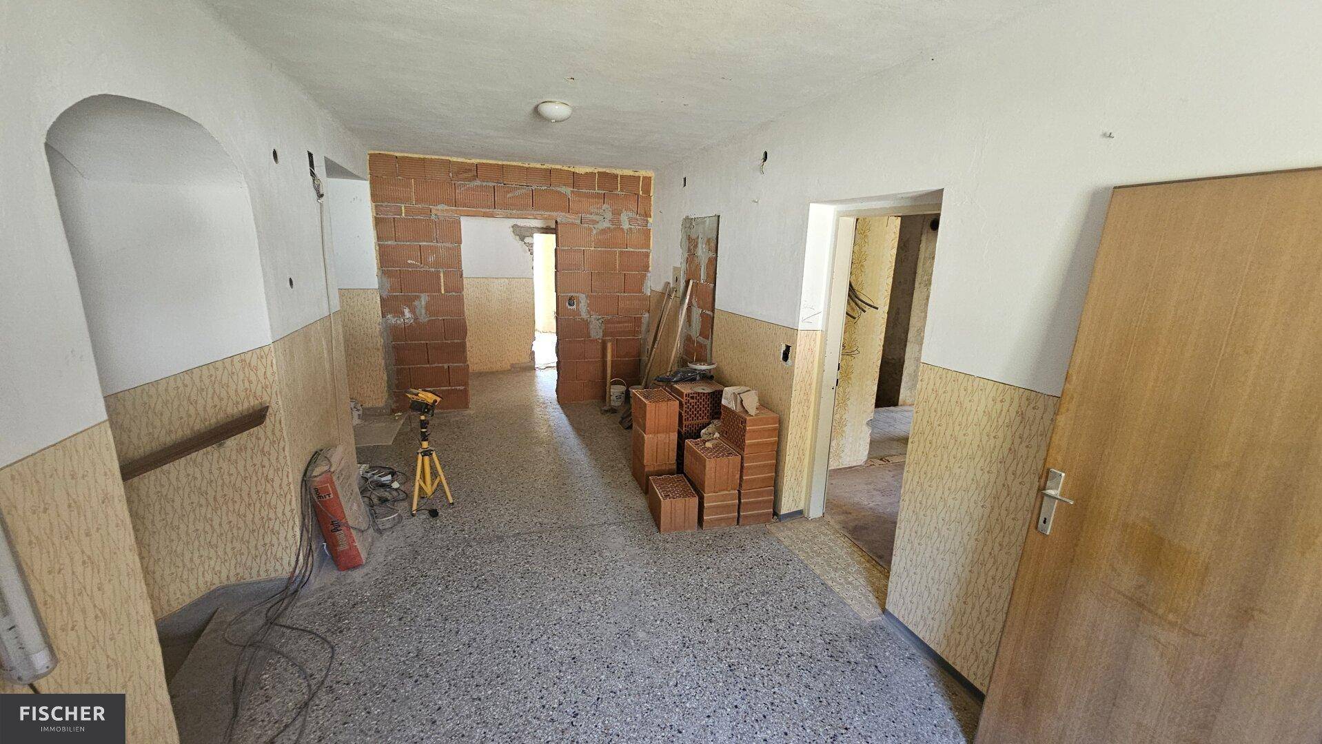 Innenraum im Rohbauzustand mit unverputzten Ziegelwänden, Terrazzoboden und Renovierungsmaterialien.