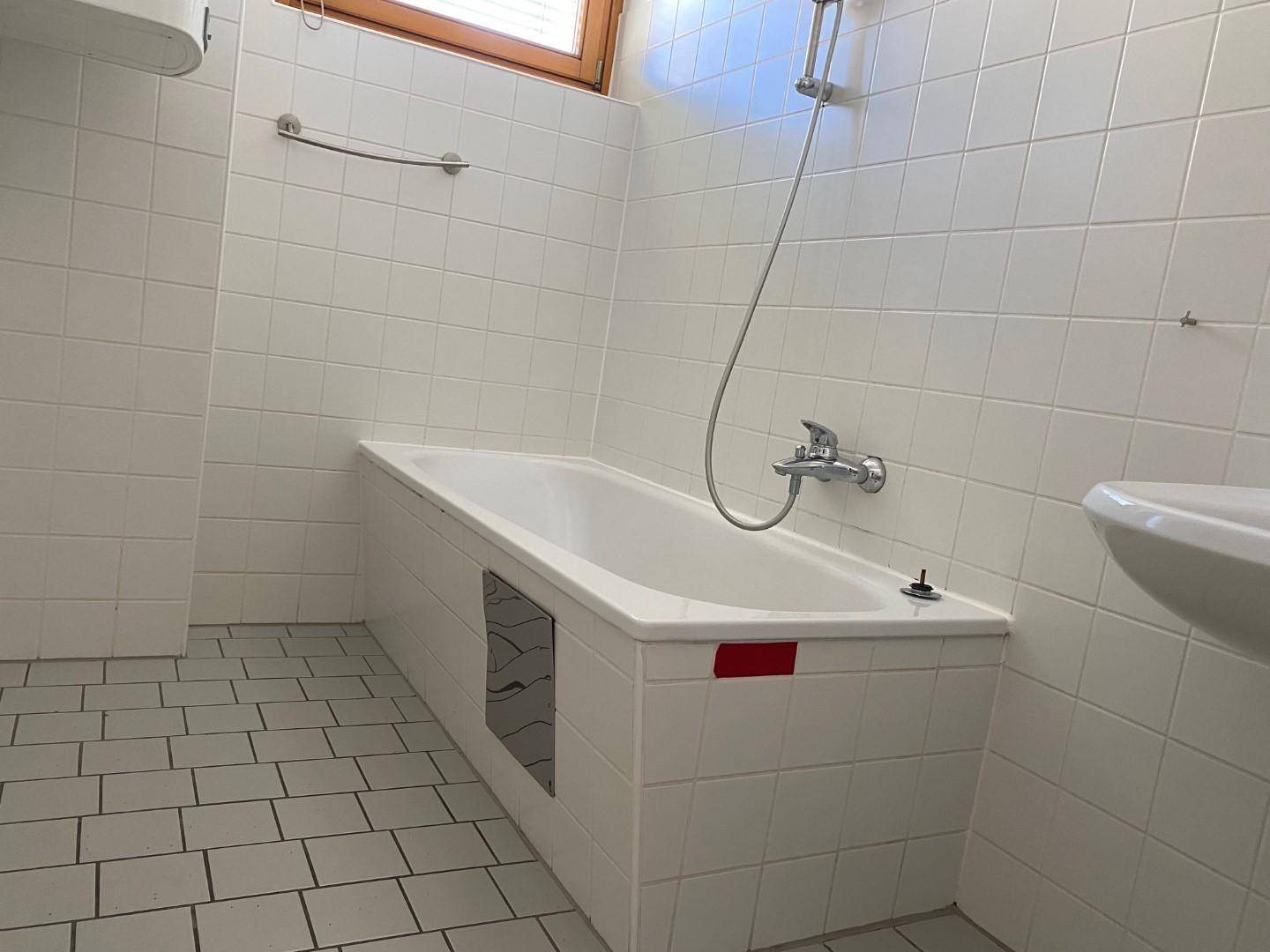 Badezimmer mit weiß gefliesten Wänden und Boden, ausgestattet mit einer Badewanne und Duscharmatur.