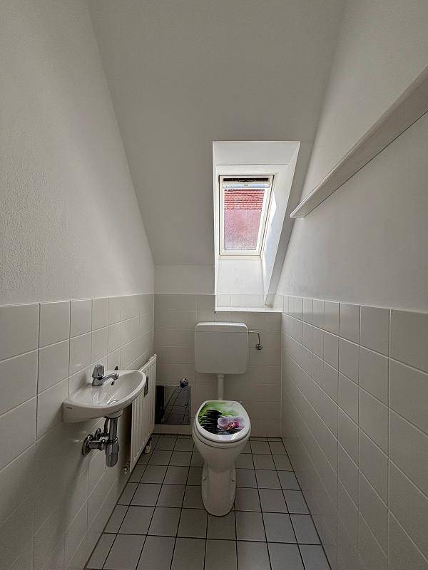 Kleines WC mit weißen Fliesen, Toilette, Handwaschbecken und einem Dachfenster unter der Dachschräge.