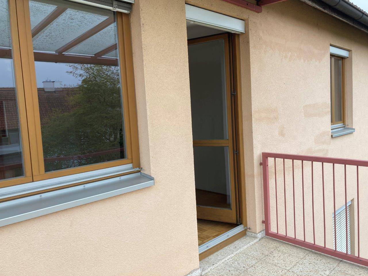 Balkonzugang mit Holztür, Fenster und rotem Metallgeländer, teilweise überdacht.