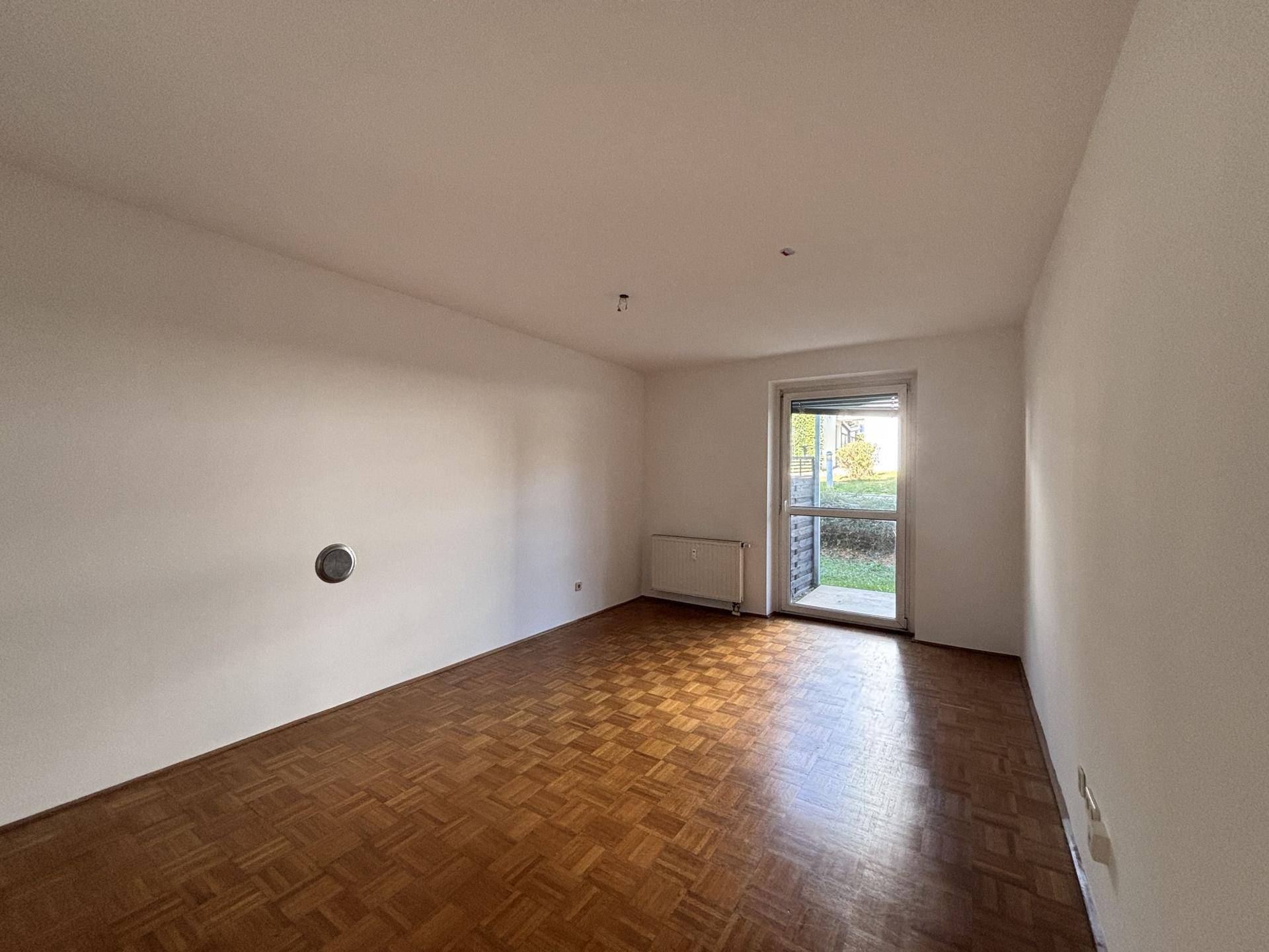 Geräumiges Wohnzimmer mit Parkettboden und Zugang zu einem Außenbereich durch eine Fenstertür.