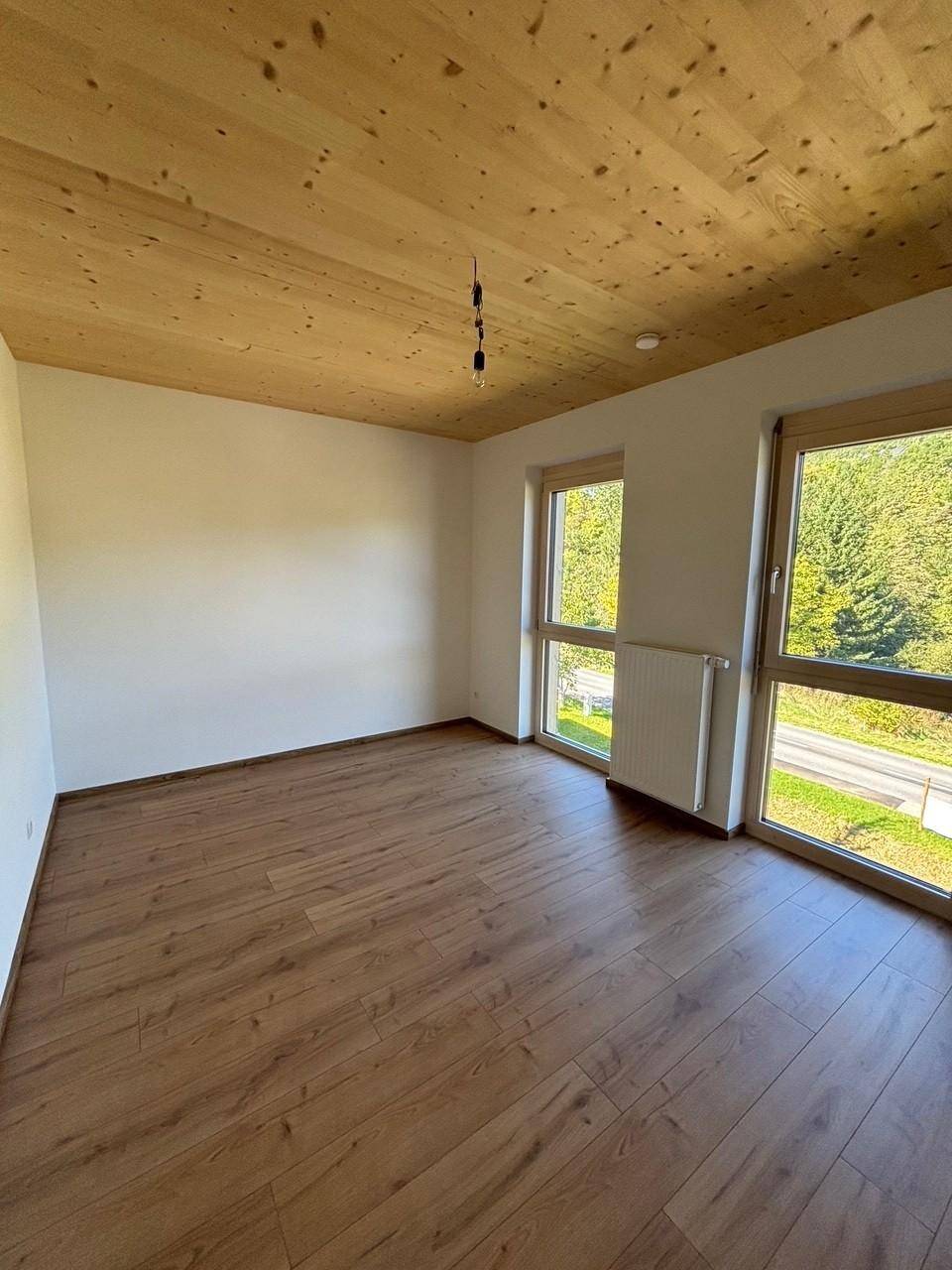 Geräumiges Zimmer mit Holzboden und Holzdecke, großen Fenstern und Blick ins Grüne.