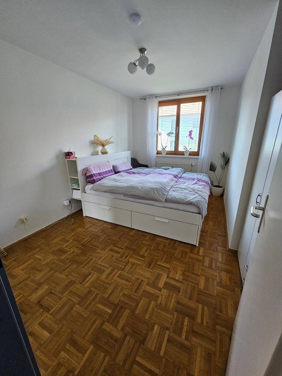 Helles Schlafzimmer mit Parkettboden, Doppelbett und Fenster mit Jalousien.