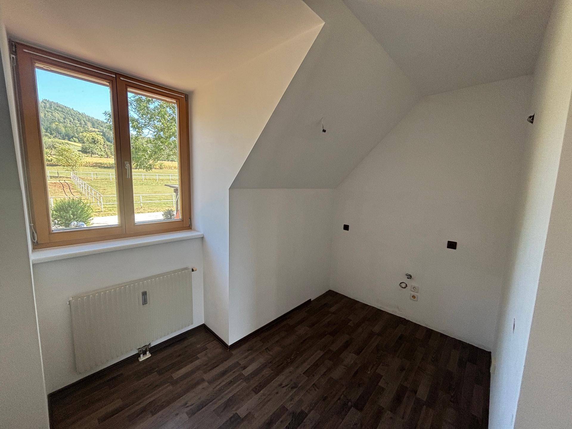 Kleiner Raum mit dunklem Laminatboden, Dachschräge und Fenster mit Blick ins Grüne.