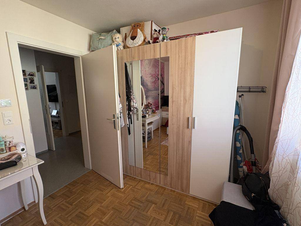 Schlafzimmer mit hellem Kleiderschrank, Spiegeltüren und Parkettboden, der zu einem weiteren Raum führt.