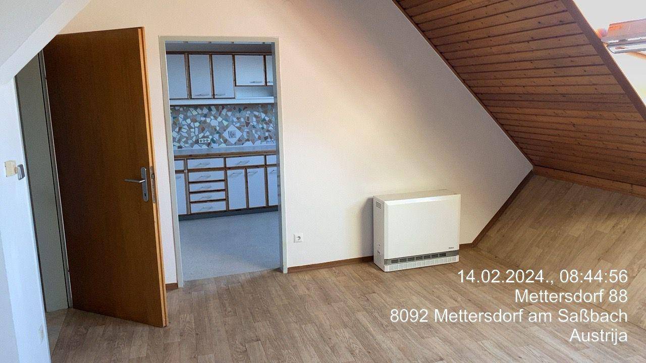 Leerer Raum mit Laminatboden, Heizkörper und Blick in die angrenzende Küche im Dachgeschoss.