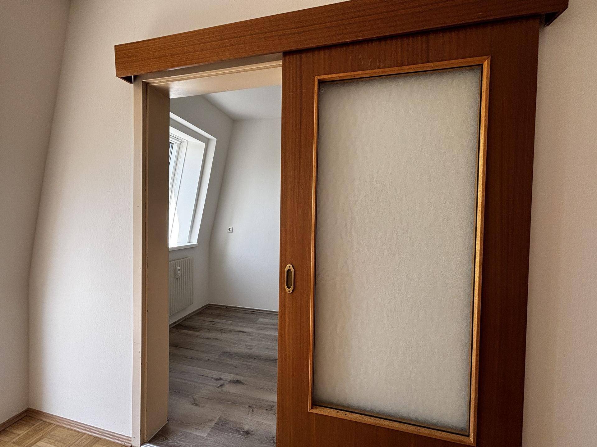 Schiebetür mit Glaseinsatz, die in einen Raum mit Dachschräge und Fenster führt.