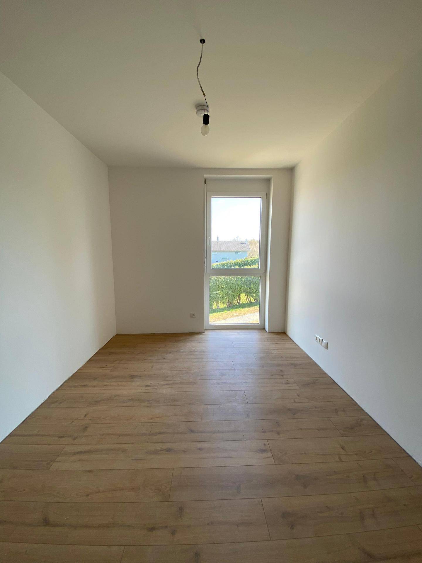 Helles, leeres Zimmer mit Laminatboden und einem Fenster mit Blick ins Grüne.