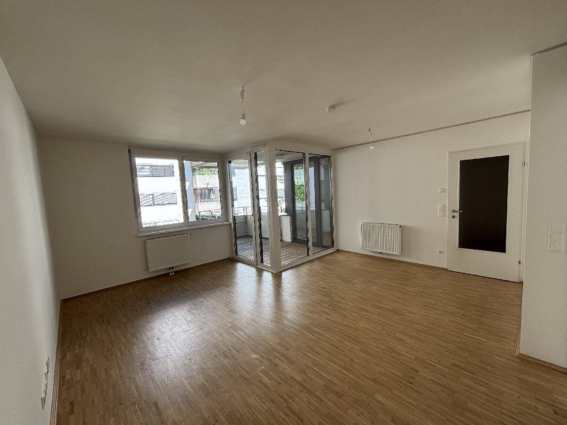 Geräumiger Wohnbereich mit hellem Holzparkettboden und viel Tageslicht durch große Fenster.