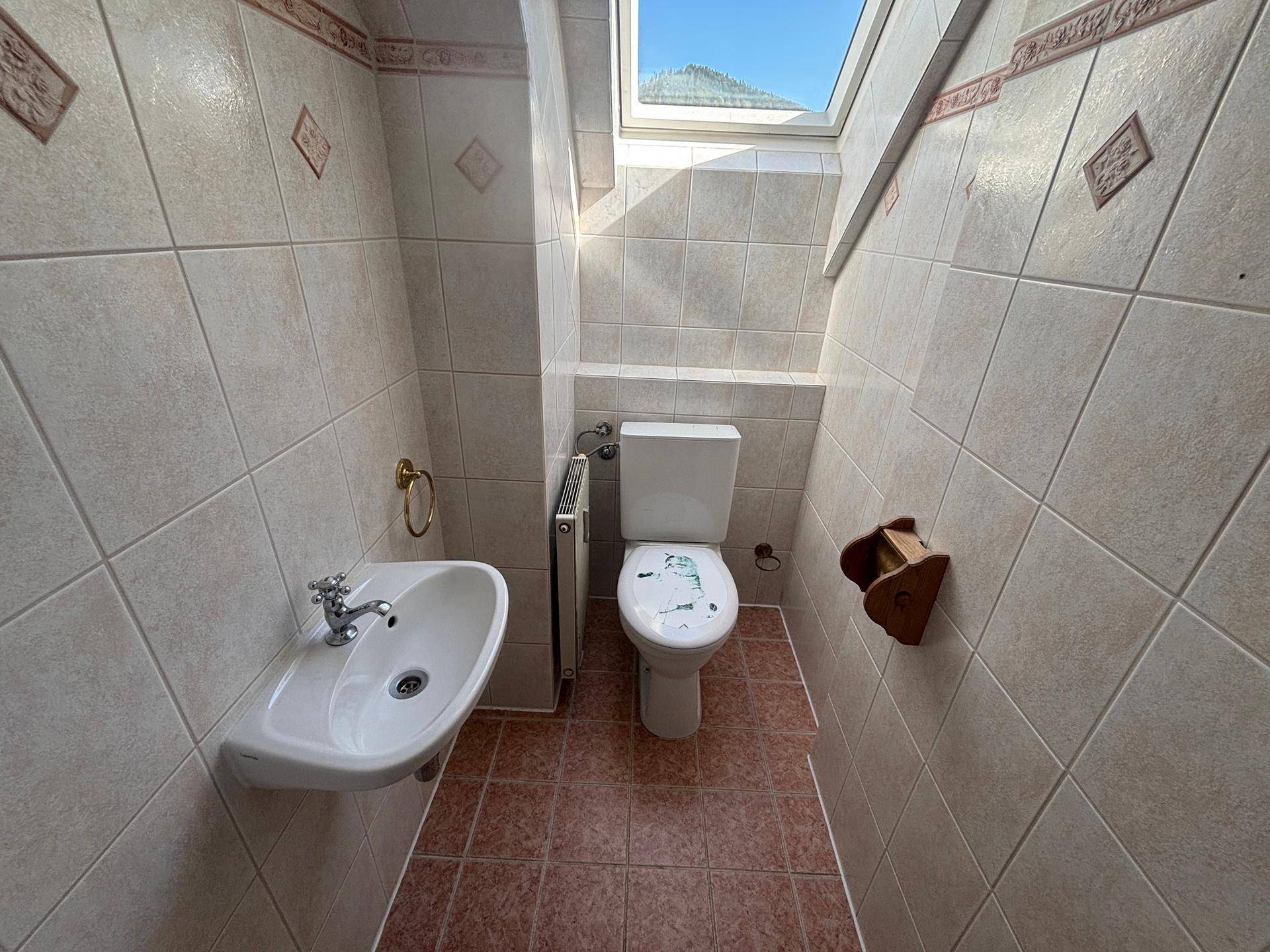 Kleines Gäste-WC mit beigefarbenen Fliesen, Toilette, Waschbecken und Dachfenster.