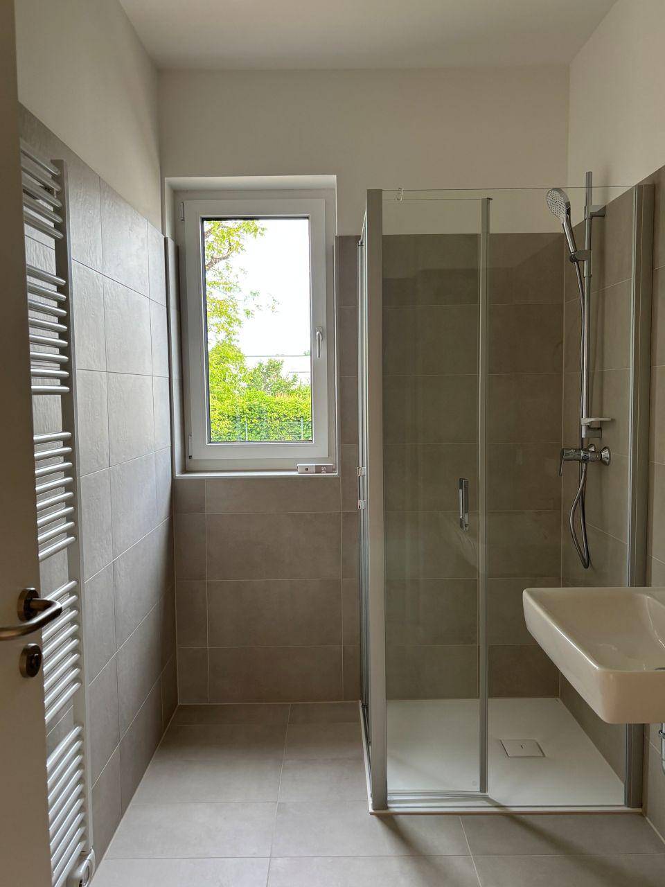 Modernes Badezimmer mit grauen Fliesen, Duschkabine und Fenster für Tageslicht.