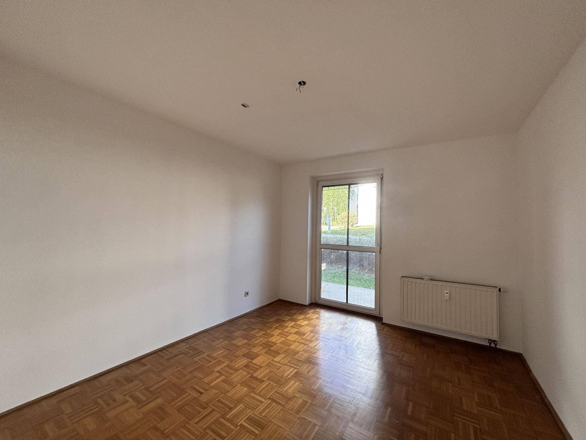 Helles Zimmer mit Parkettboden und einer Fenstertür, die zu einem Außenbereich führt.