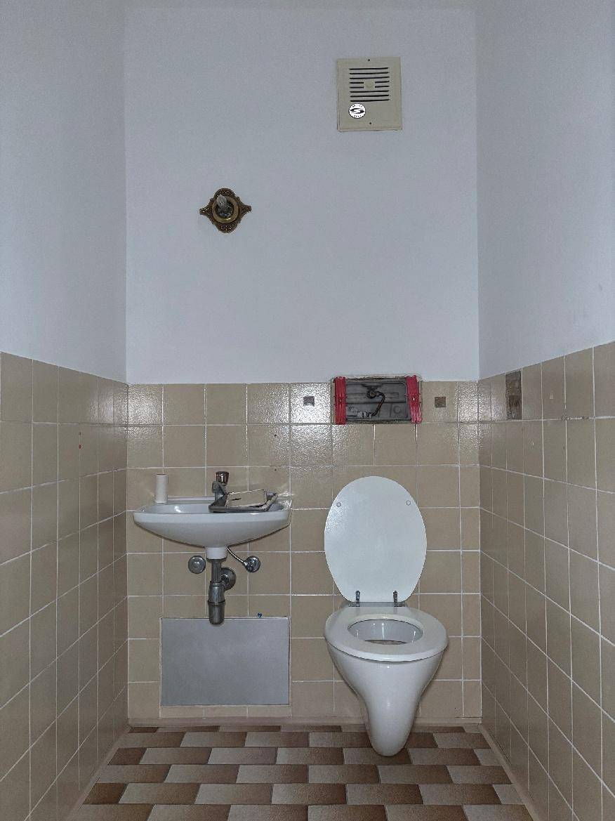 Separates WC mit Toilette und kleinem Waschbecken, gefliest in beigen Farbtönen.