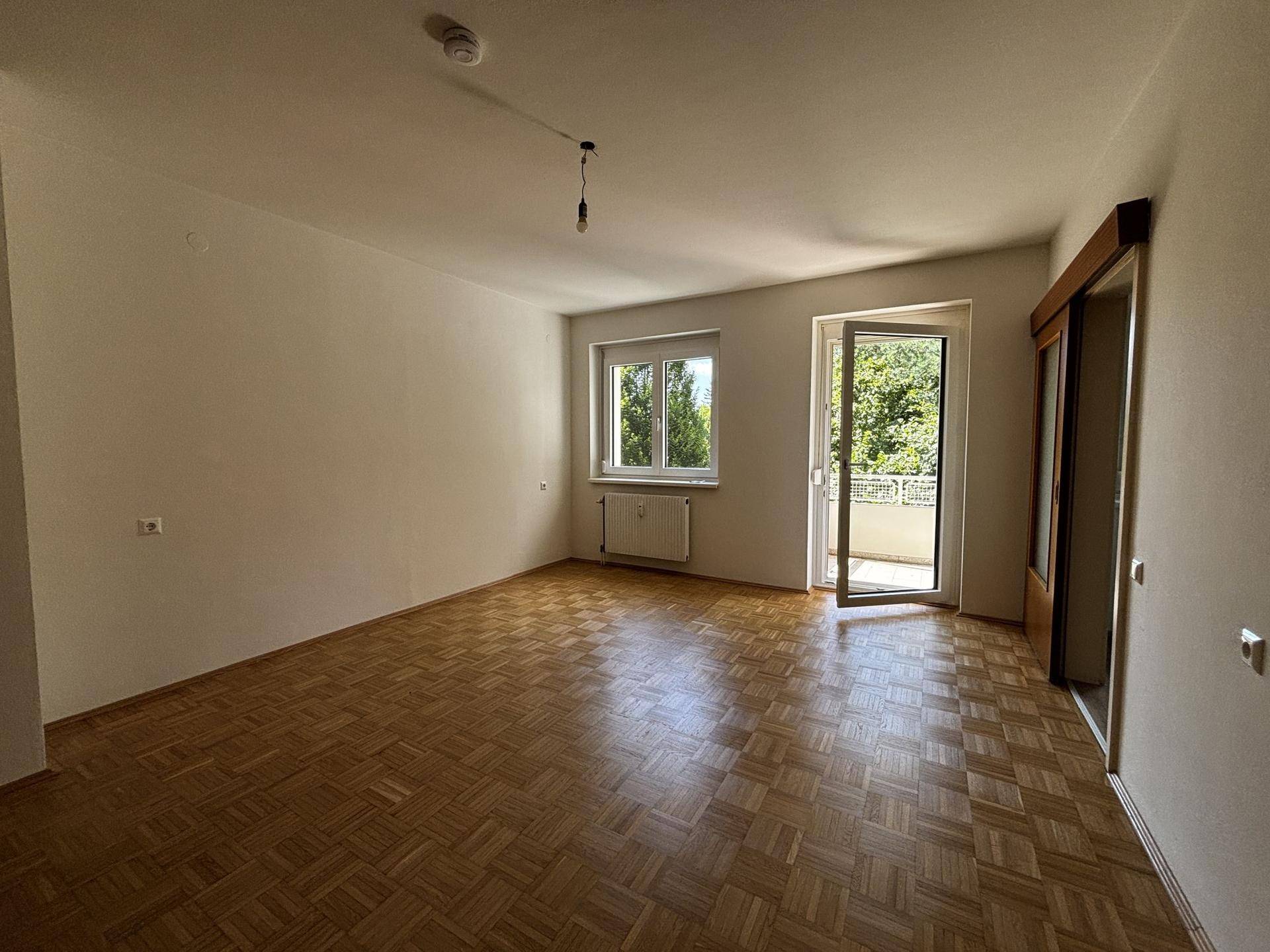 Helles Wohnzimmer mit Fischgrätparkett, großem Fenster und Zugang zu einem sonnigen Balkon.
