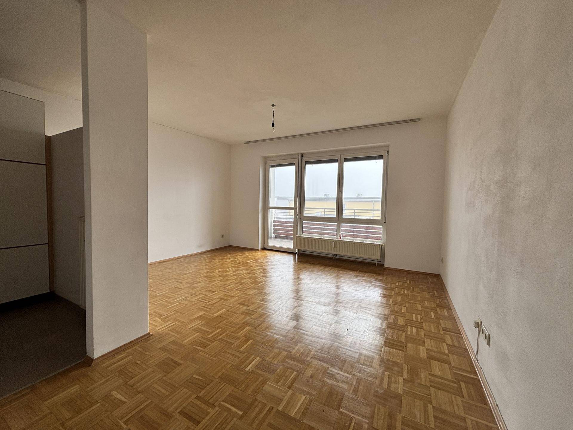 Geräumiges Wohnzimmer mit Parkettboden und großen Fenstern, die Zugang zum Balkon bieten.