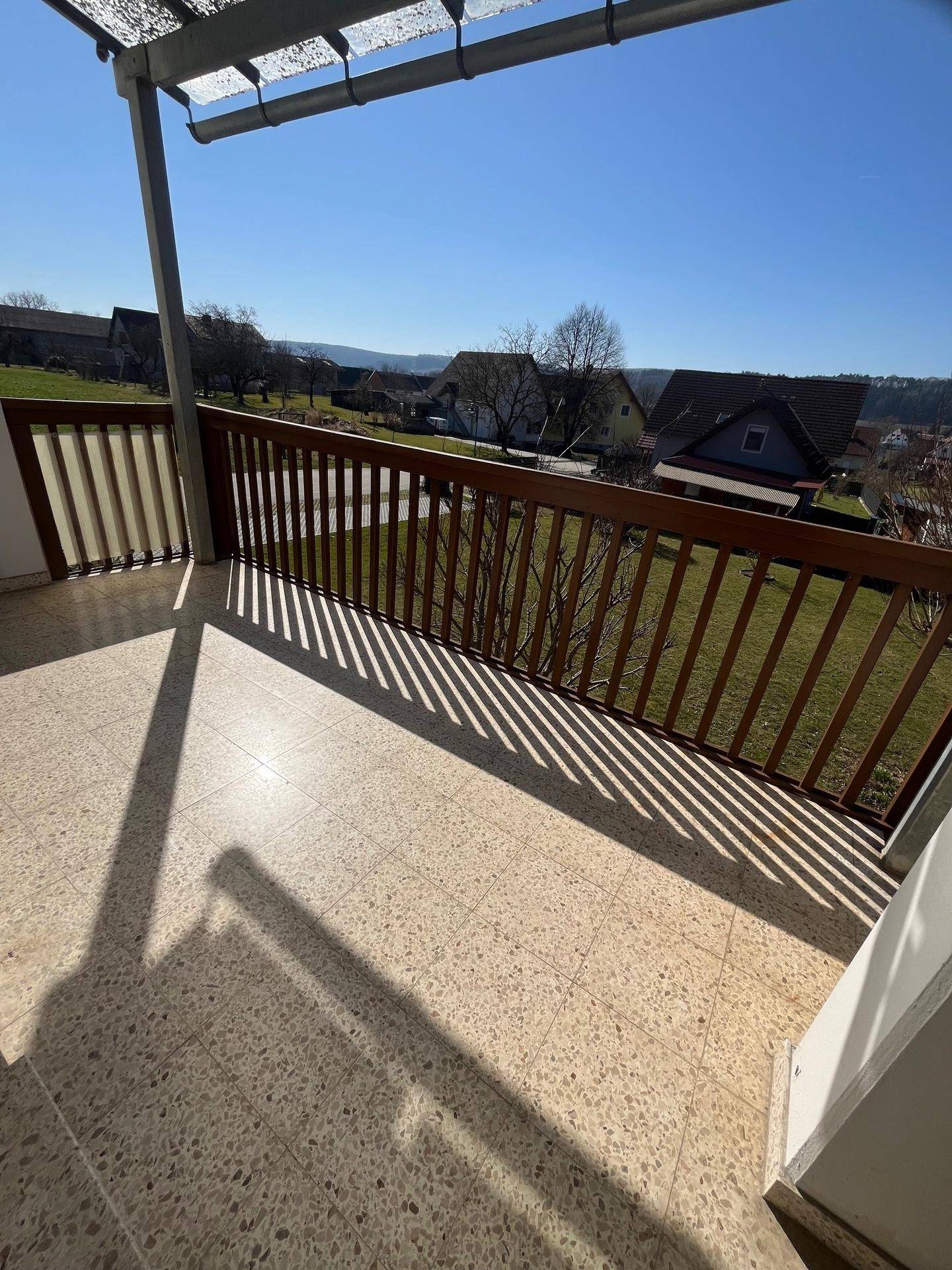Sonniger Balkon mit Fliesenboden, Holzgeländer und weitem Blick über die grüne Landschaft.