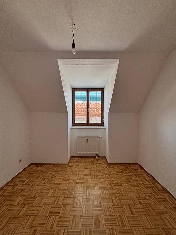 Helles Zimmer im Dachgeschoss mit Parkettboden, schrägen Wänden und einem Fenster mit Blick auf die Dächer.