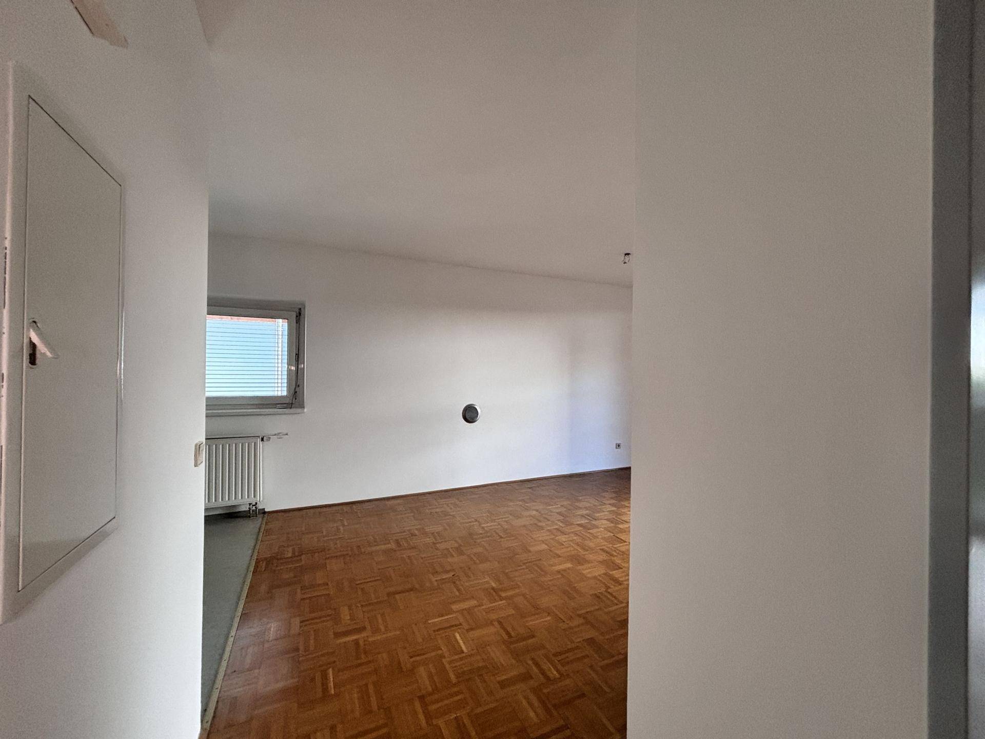 Leeres Wohnzimmer mit Parkettboden und einem Fenster, das Tageslicht hereinlässt.
