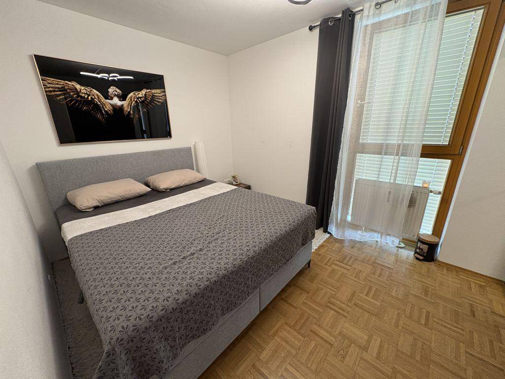 Helles Schlafzimmer mit Doppelbett, Parkettboden und großem Fenster mit Vorhängen.