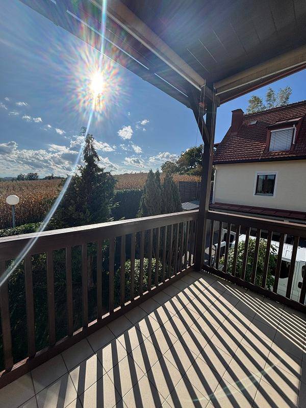 Sonniger Balkon mit Holzgeländer und Blick auf die umliegende Natur sowie das angrenzende Gebäude.