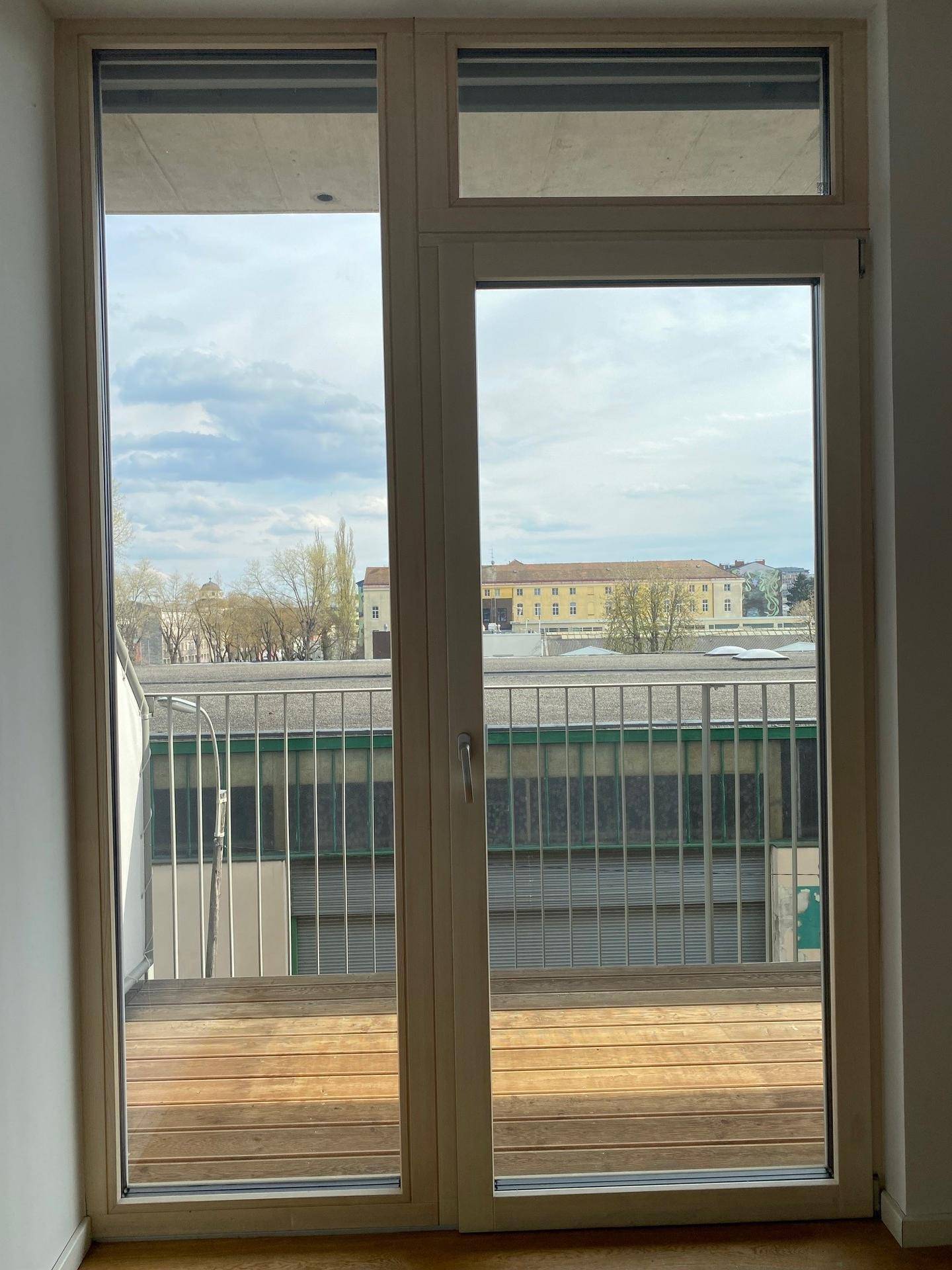 Helles Zimmer mit Zugang zu einem Balkon mit Holzbelag und Blick auf die umliegende Stadt.