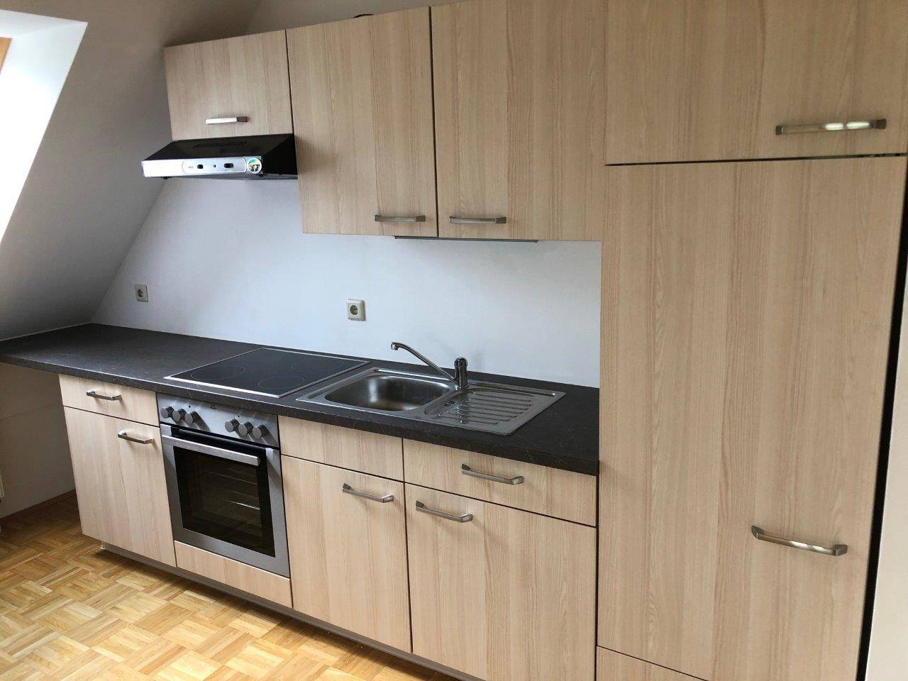 Kompakte Einbauküche mit hellen Holzfronten, Kochfeld und Backofen unter Dachschräge.