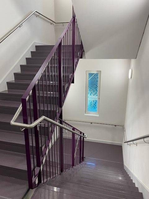 Modernes Treppenhaus mit lila Geländer, grauen Stufen und einem schmalen, gemusterten Fenster.