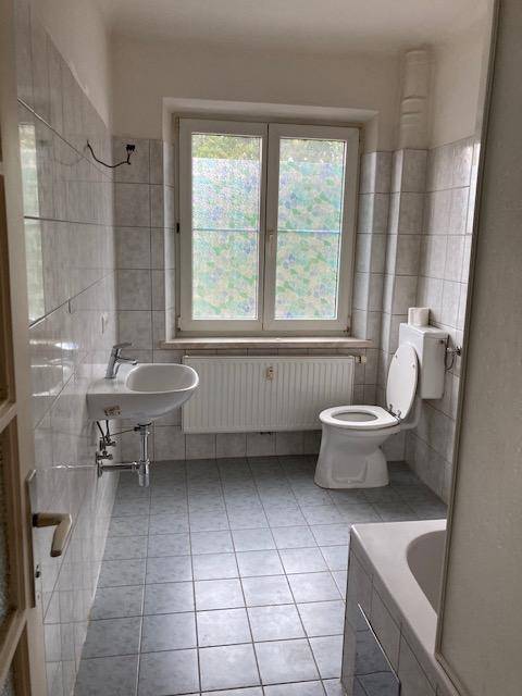 Helles Badezimmer mit Fenster, WC, Waschbecken und hellgrauen Fliesen.