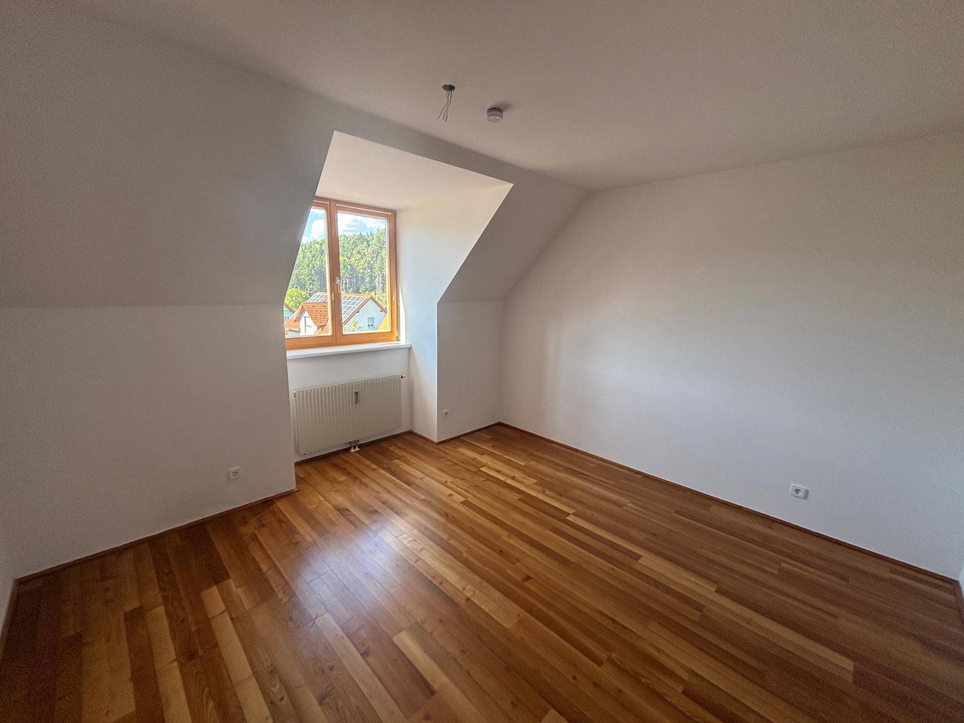 Helles Schlafzimmer mit Holzparkett, Dachschrägen und Fenster mit Blick auf die Umgebung.