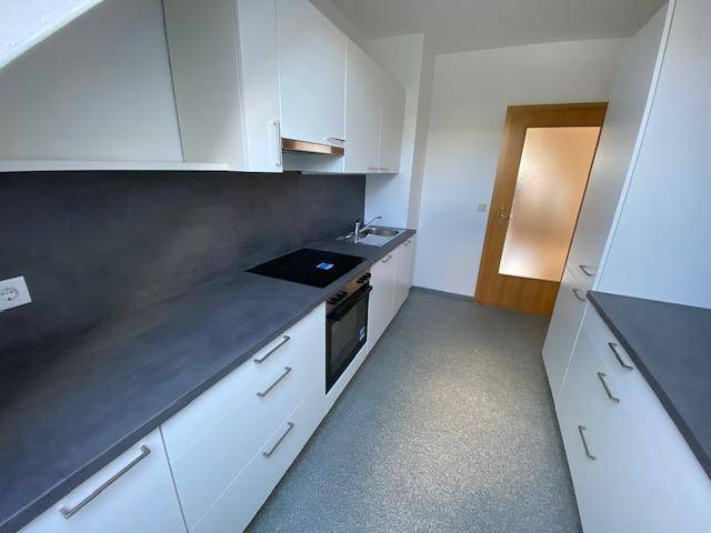 Geräumige Küche mit durchgehender Arbeitsplatte, weißen Unterschränken und einer modernen Holztür.