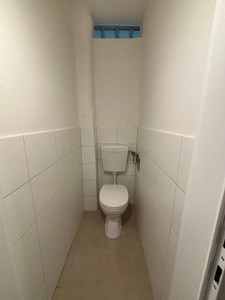 Separates WC mit Toilette und halbhohen weißen Fliesen, beleuchtet durch ein Oberlicht.