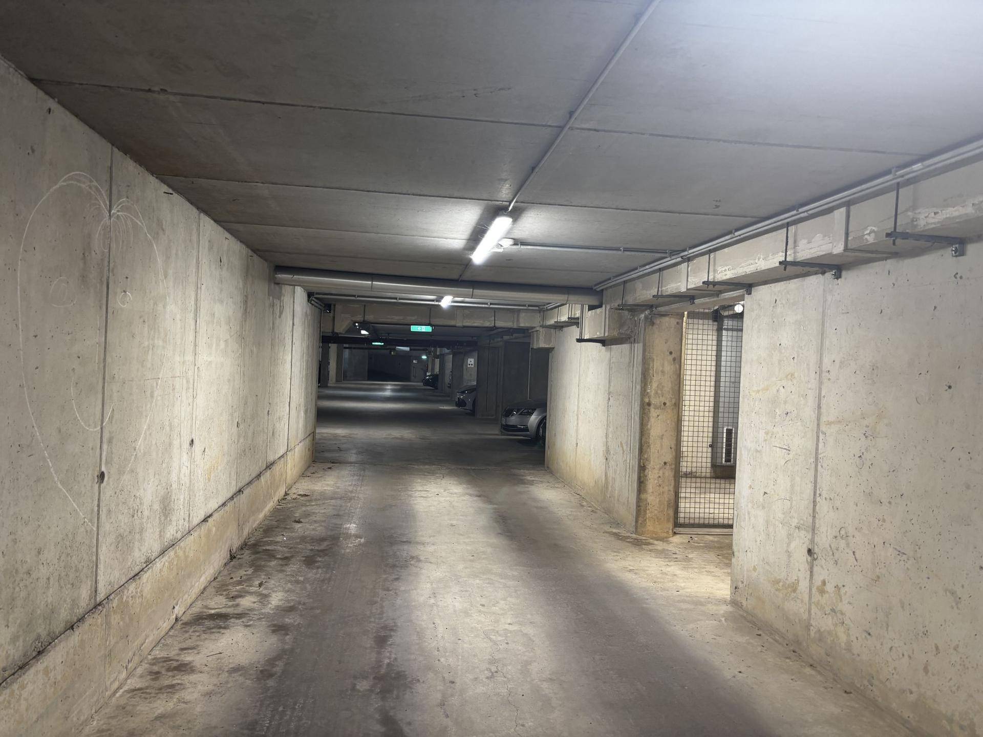 Langer, betonierter Gang einer Tiefgarage mit Beleuchtung und sichtbaren Installationen an der Decke.