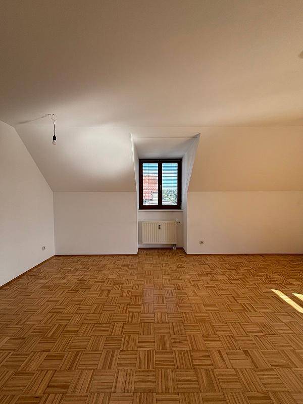 Geräumiges Zimmer im Dachgeschoss mit Parkettboden, schrägen Wänden und einem Fenster mit Blick auf die Dächer.