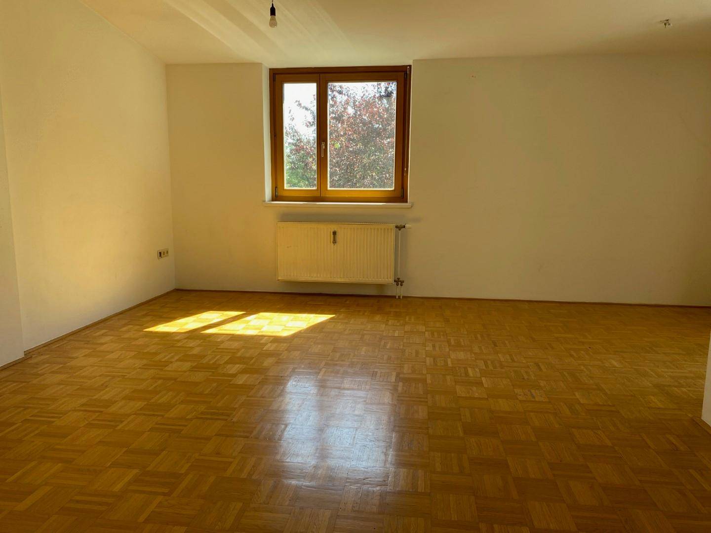 Geräumiges Wohnzimmer mit hellem Parkettboden, großem Fenster und viel Tageslicht.