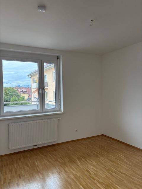 Leeres Schlafzimmer mit Parkettboden, weiß gestrichenen Wänden und einem Fenster mit Ausblick.