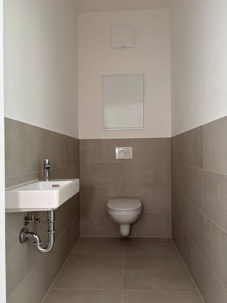 Modernes Gäste-WC mit wandhängender Toilette, kleinem Waschbecken und grauen Fliesen.