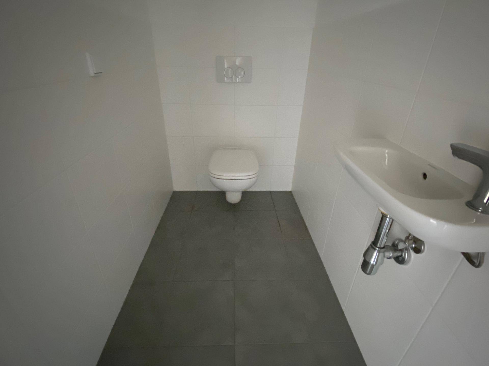 Separates WC mit Wand-WC, kleinem Waschbecken und grauen Bodenfliesen.
