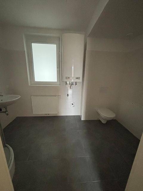 Helles Badezimmer mit Toilette, Waschbecken, Fenster und Warmwasserbereiter an der Wand.