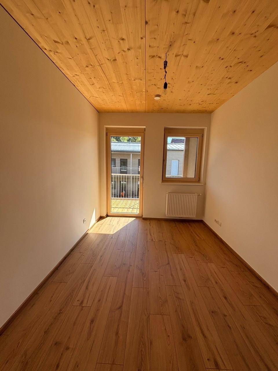 Helles Zimmer mit Holzboden und Holzdecke, Balkonzugang und einem zusätzlichen Fenster.