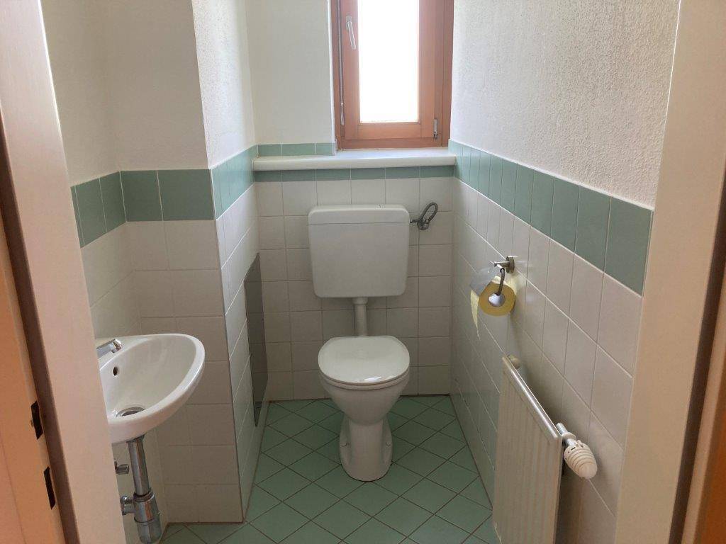 Separate Gästetoilette mit Fenster, WC und kleinem Handwaschbecken in hellen Fliesen.