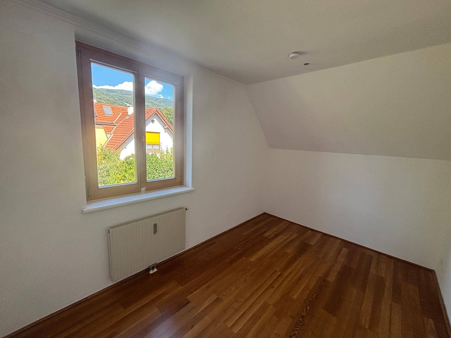 Gemütliches Schlafzimmer mit Holzparkett, Dachschräge und Fenster mit Ausblick auf Häuser und Bäume.